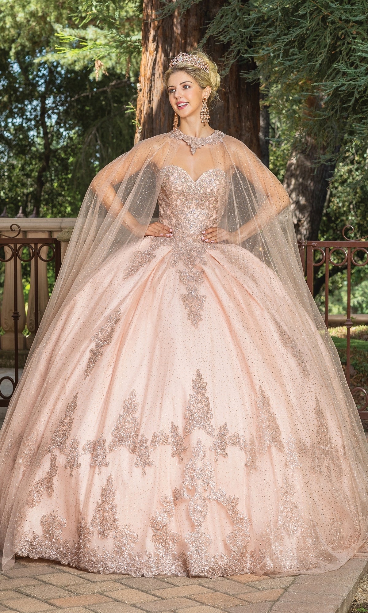 A woman stands outdoors in the Quinceanera Dress 1771 By Dancing Queen, a light pink A-line ball gown with a strapless sweetheart neckline, sparkling sheer cape, jeweled tiara, and statement earrings. Trees and a fence are behind her.